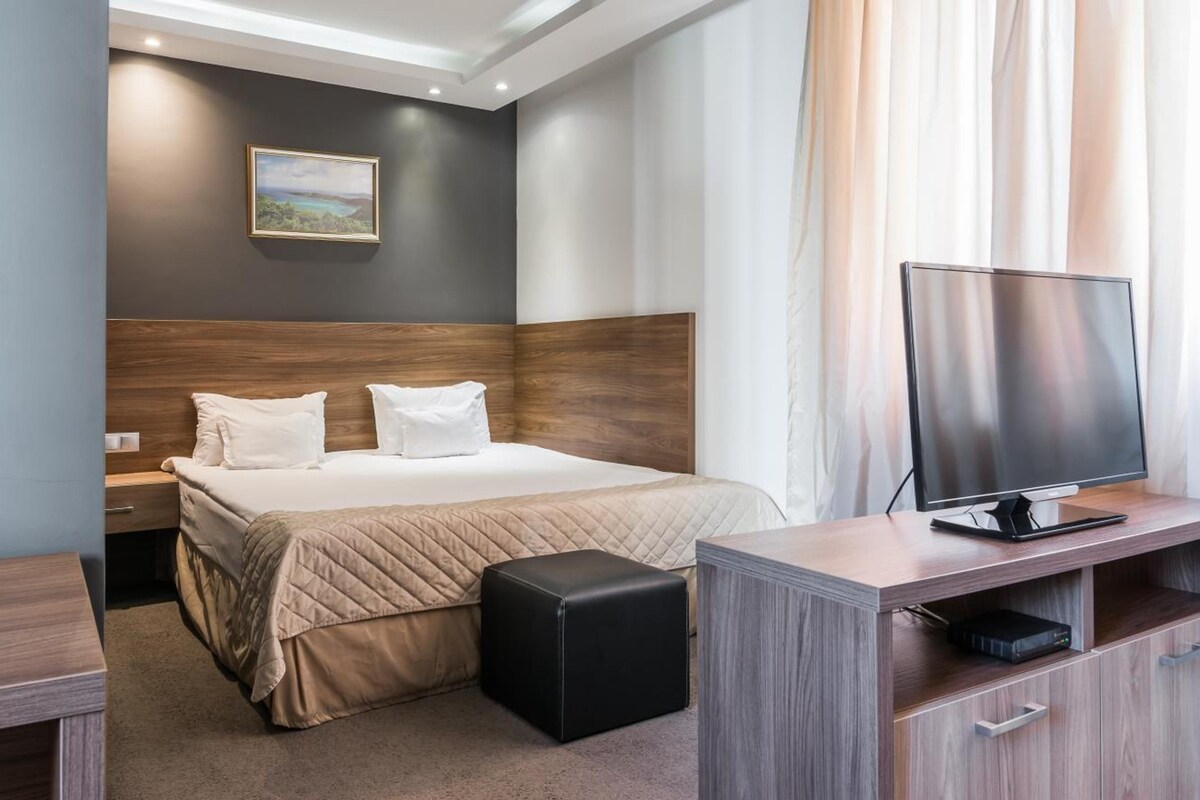 A hotel room with a bed, a TV, a desk, and a small ottoman.