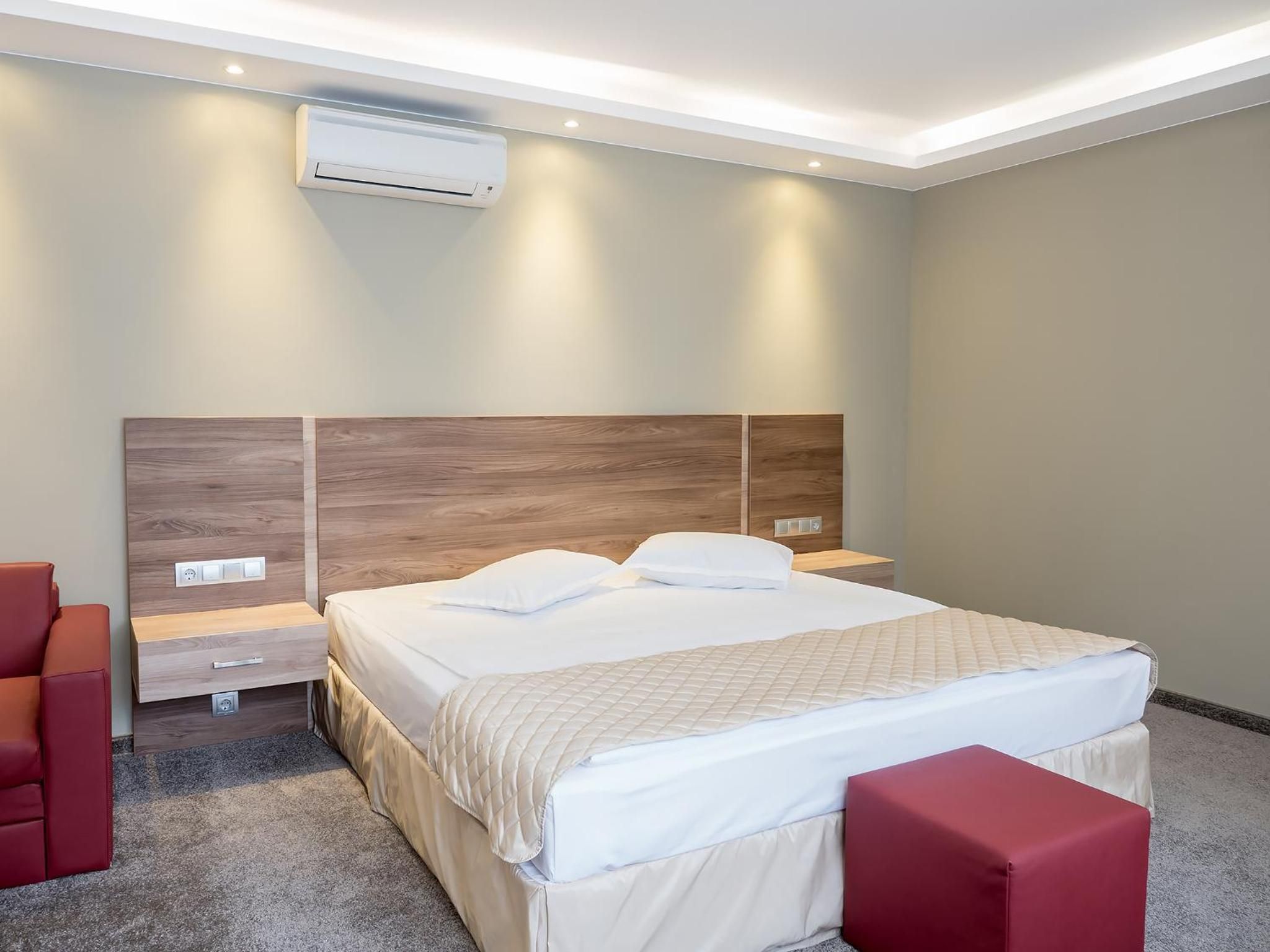 A hotel room with a bed, bedside tables, an air conditioning unit, and a red chair.