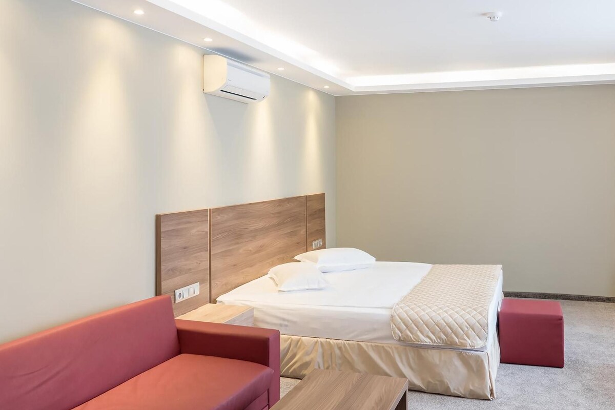 A hotel room with a bed, a red sofa, a wooden table, and a wall-mounted air conditioning unit.