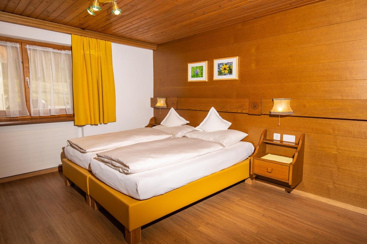 Una cama doble con sábanas blancas, cabecera de madera, dos lámparas de noche, mesita con cajón y una ventana con cortinas amarillas.