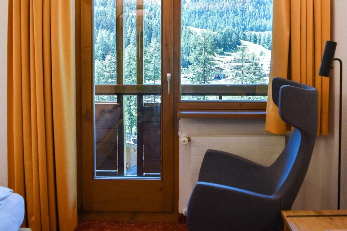Habitación de hotel con cama, dos sillas, escritorio, ventana con cortinas y vistas a zona boscosa.
