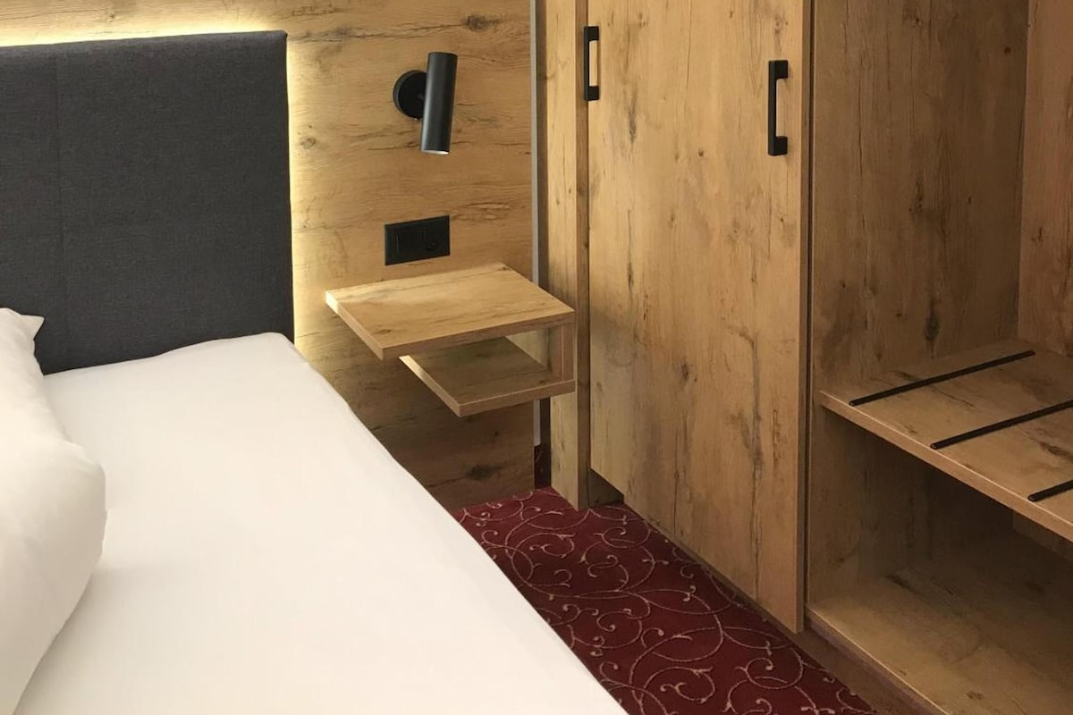 Una habitación de hotel con una cama, un armario de madera, una mesita de noche y una lámpara de pared.