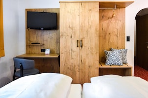 A hotel room with a wooden wardrobe, a television mounted on the wall, a desk with a chair, and two beds with pillows.