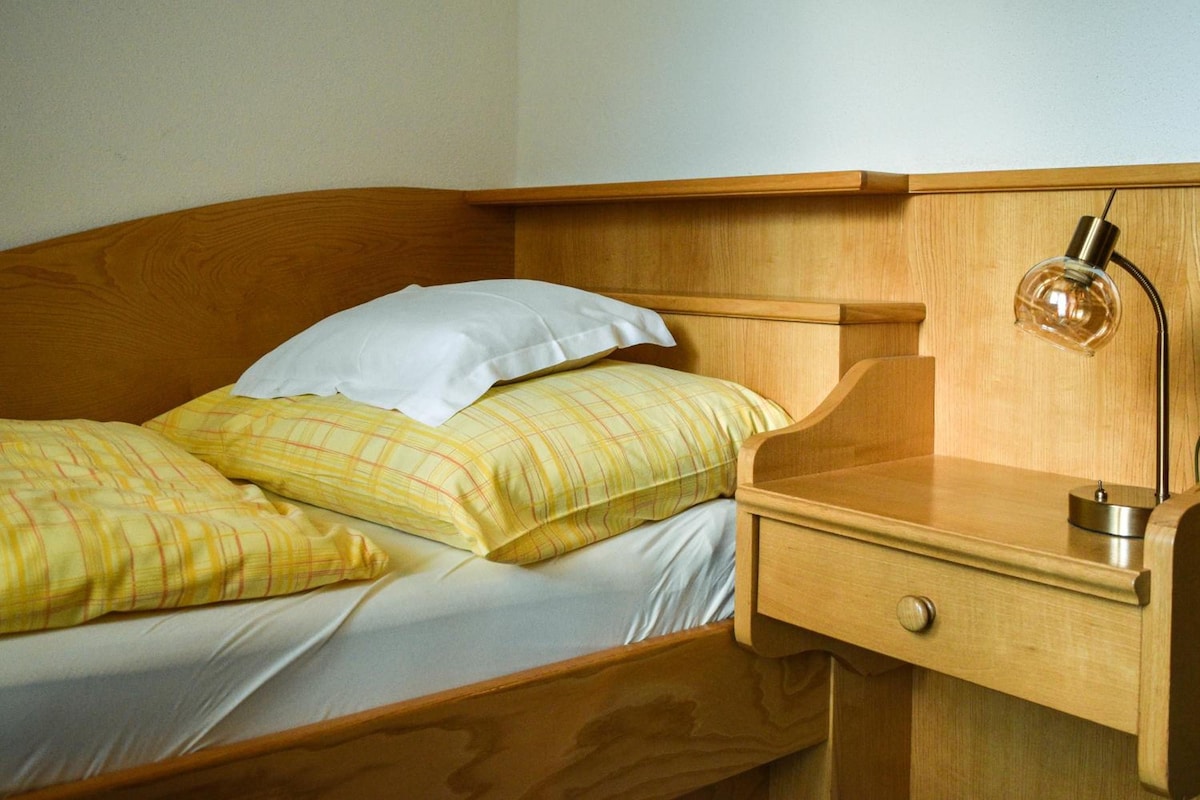 Una cama de madera con dos almohadas, una mesita de noche con una lámpara y un cajón.