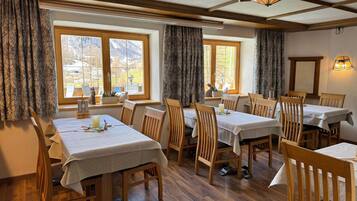 A dining area with multiple tables and chairs, large windows with curtains, and a wooden ceiling.