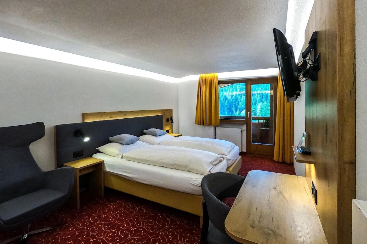 Habitación de hotel con cama, escritorio, silla, televisor y ventana con cortinas.