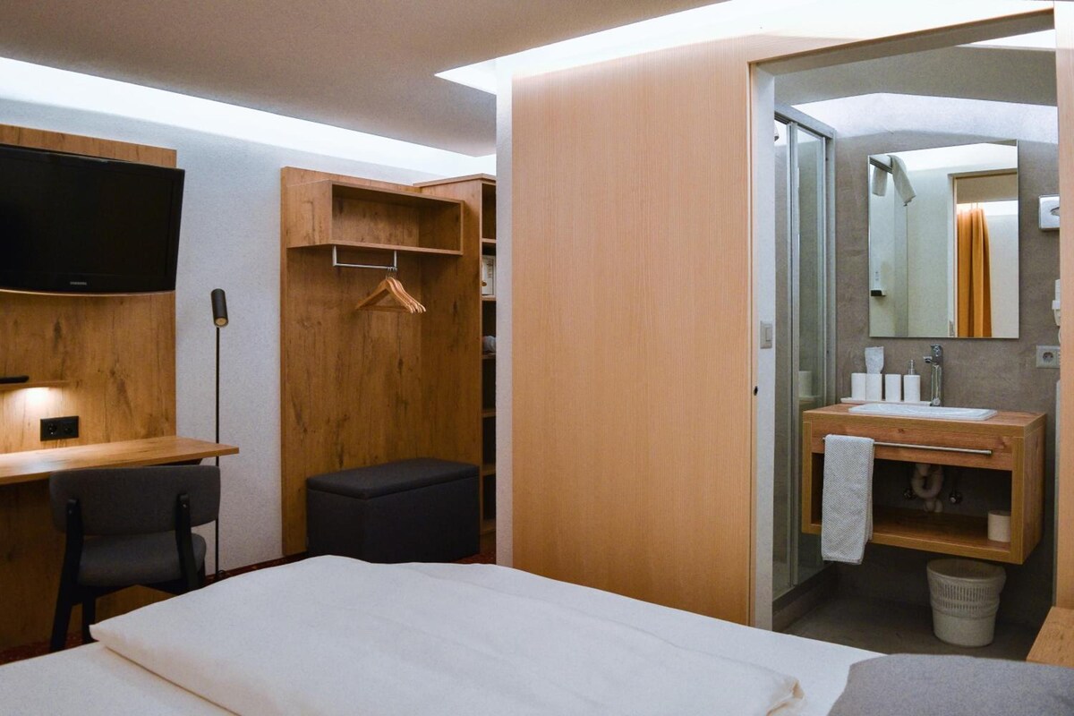 Una habitación de hotel con cama, escritorio, silla, armario de madera, perchero y baño con lavabo y espejo.