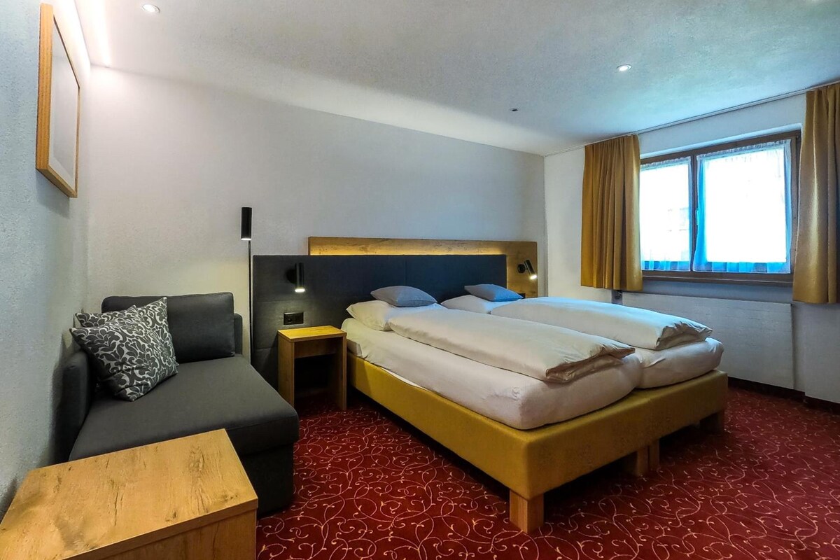 Habitación de hotel con cama doble, sofá, mesita de noche y ventana con cortinas.