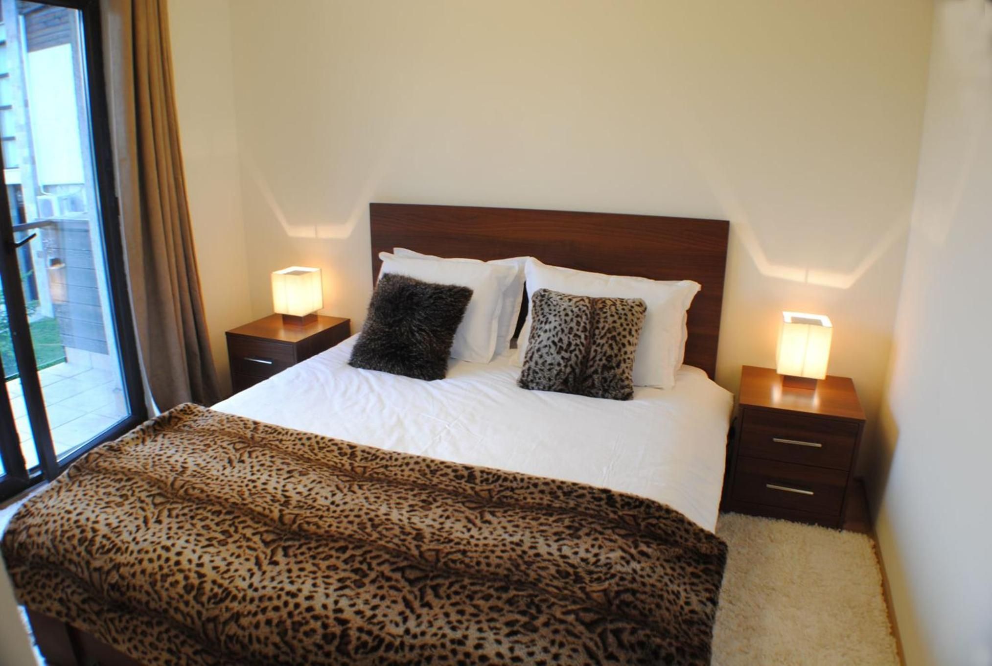 A hotel room with a bed, two bedside tables with lamps, a window with curtains, and a leopard print throw blanket.