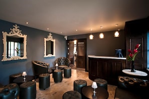 A room with a dark blue wall, two ornate mirrors, a leather armchair, a round table with a lamp, and a bar area with a vase of flowers. - The Bull At Great Totham Limited (Maldon)