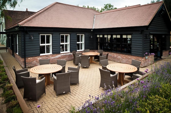 A building with a dark exterior, a tiled roof, and a patio area with wicker furniture and a round table. - The Bull At Great Totham Limited (Maldon)