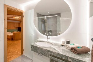 A modern bathroom with a large mirror, a granite countertop, a shower, and a wooden door leading to a bedroom. - Pension Rudolph (Gaschurn)