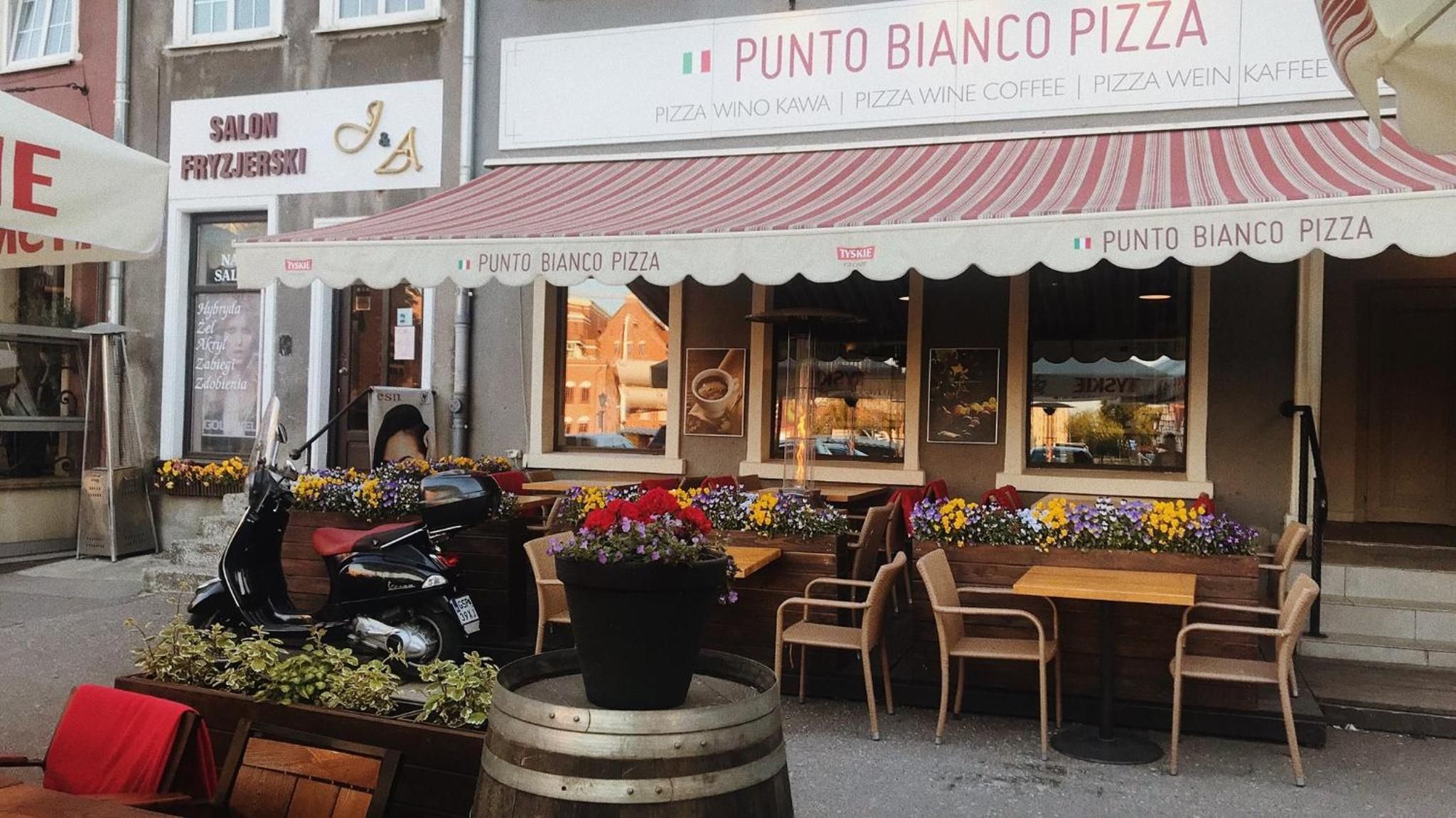An outdoor seating area with tables and chairs, a motorcycle parked outside, and a storefront with signage for Punto Bianco Pizza.
