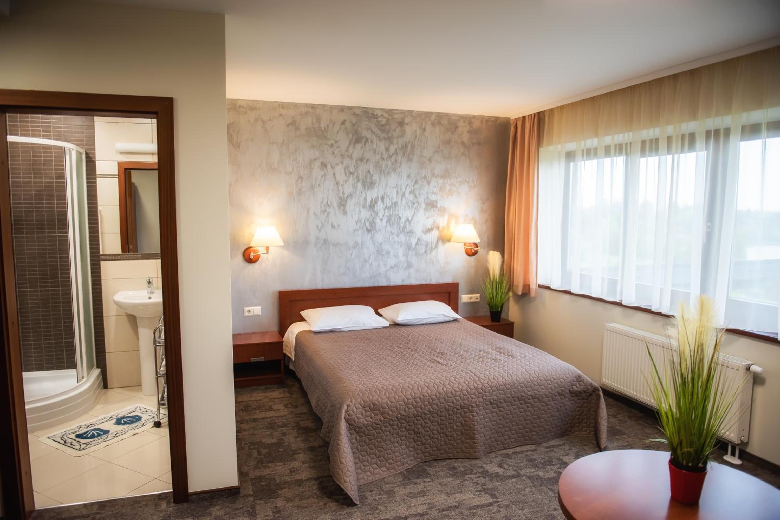 A hotel room with a bed, bedside lamps, a window with curtains, a radiator, and a bathroom with a shower and sink.