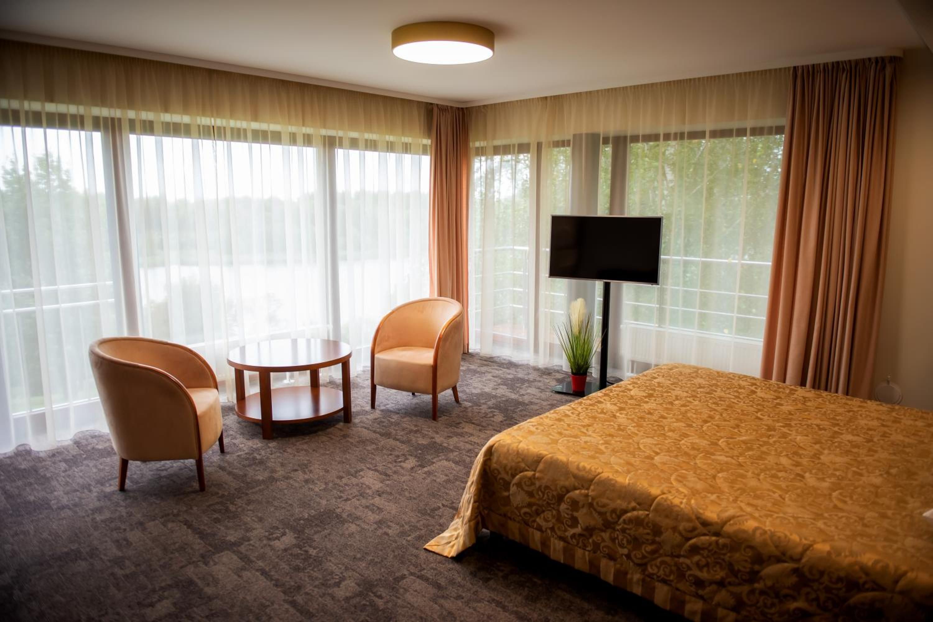 Een hotelkamer met een bed, twee fauteuils, een klein rond tafeltje, een televisie en grote ramen met gordijnen.