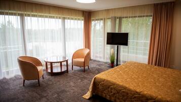 Een hotelkamer met een bed, twee fauteuils, een klein rond tafeltje, een televisie en grote ramen met gordijnen.