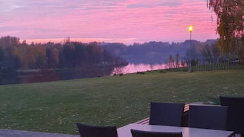 An outdoor seating area with tables and chairs, overlooking a body of water and trees during sunset.