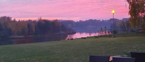 An outdoor seating area with tables and chairs, overlooking a body of water and trees during sunset.