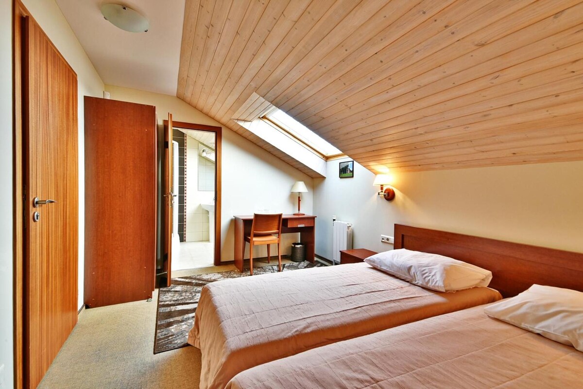 A bedroom with two beds, a desk, and a chair. There is a skylight above the desk and a door leading to another room.