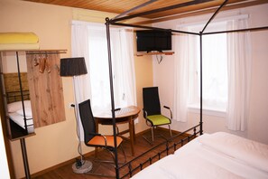 Quarto com cama, escrivaninha, duas cadeiras, televisão e janela com cortinas.