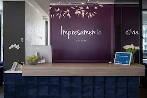 Een receptiebalie met een paarse achtergrond, een bord met de tekst 'Impresamente Art Hotel' en een lijst met een welkomstboodschap en een beoordeling van 9,7.