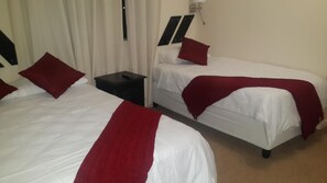 Two single beds with white bedding and red accents, a small bedside table, and a wall-mounted light fixture.