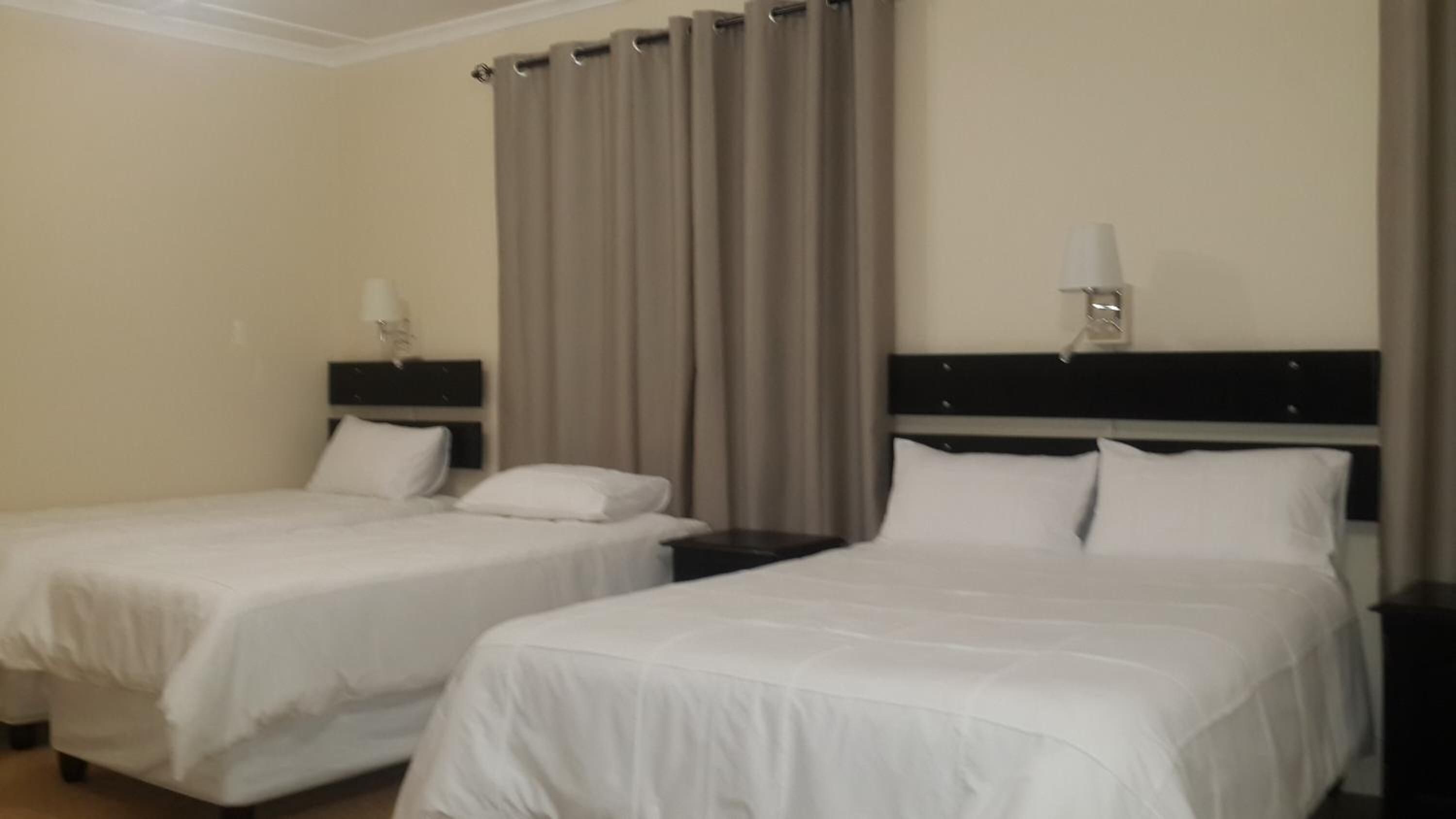 Habitación de hotel con dos camas, una mesita de noche con lámpara y una ventana con cortinas.