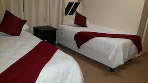 Two single beds with white linens and red accents, a small black nightstand, and a wall-mounted light fixture.