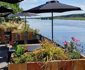 An outdoor seating area with wooden planters containing various plants and flowers, overlooking a calm river. - Baron Frits (Kessel)