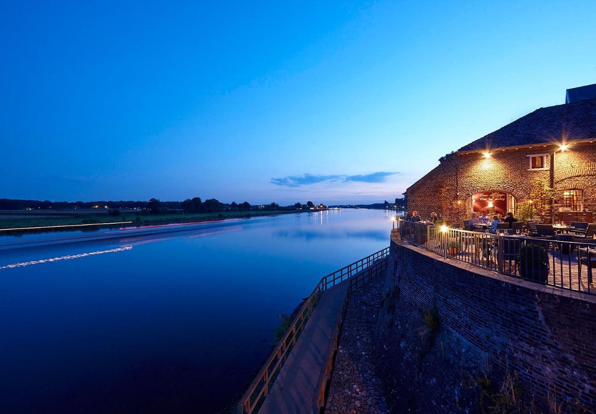 Une vue sur la rivière, avec un bâtiment en briques doté d’une terrasse, d’un mur en pierre et d’une étendue d’eau paisible.
