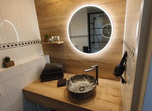 A bathroom with a wooden vanity, a round mirror with a light, a sink with a marble basin, and a towel rack. - Baron Frits (Kessel)