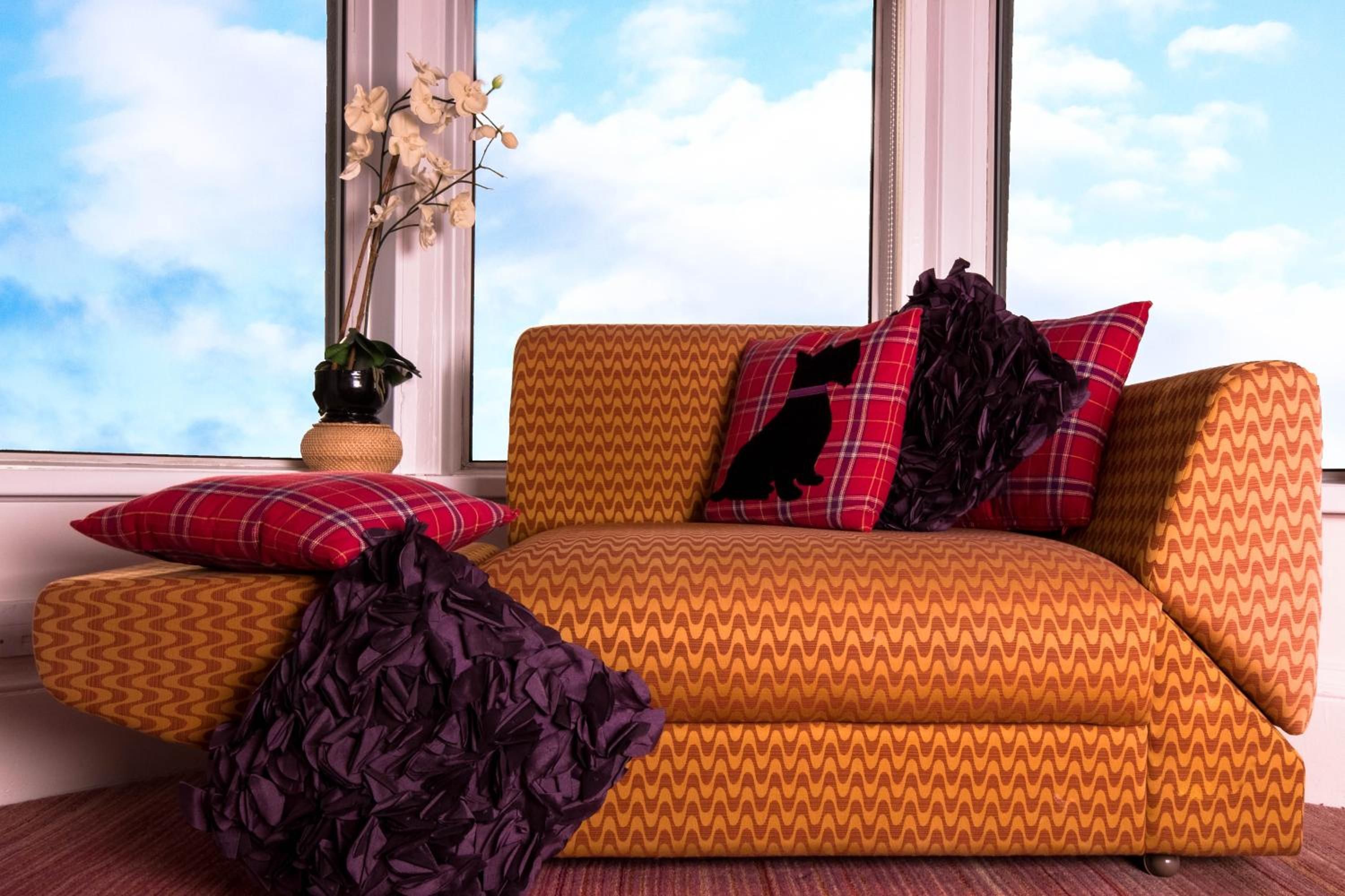 A room with a sofa, pillows, a vase with flowers, and a window with a view of the sky.