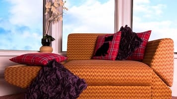 A room with a sofa, pillows, a vase with flowers, and a window with a view of the sky.