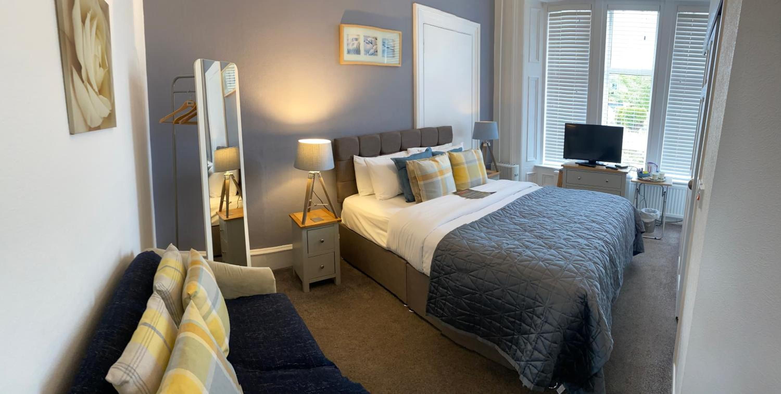 A bedroom with a bed, bedside tables, a mirror, a TV, and a window with blinds.