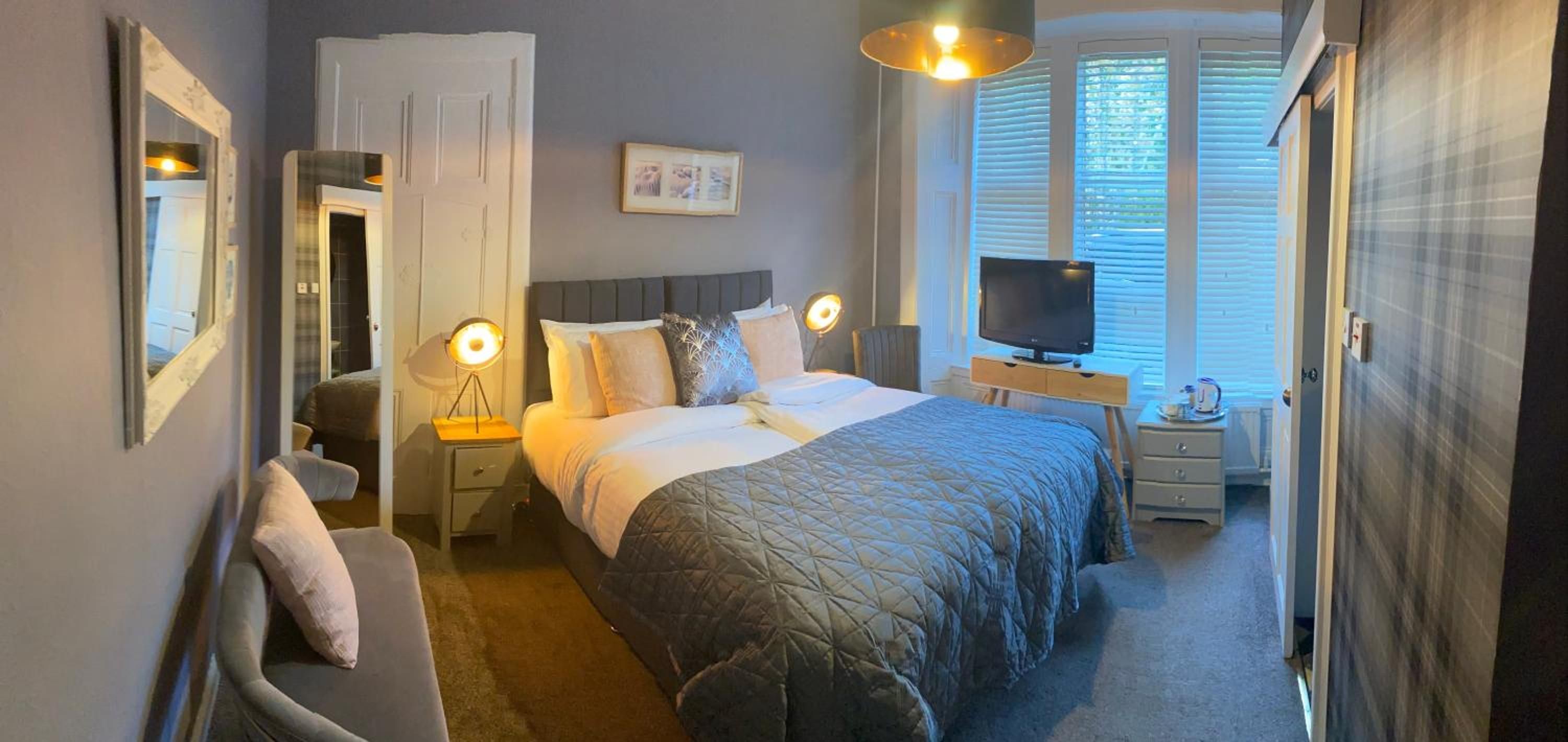A hotel room with a bed, bedside tables, a TV, and a window with blinds.
