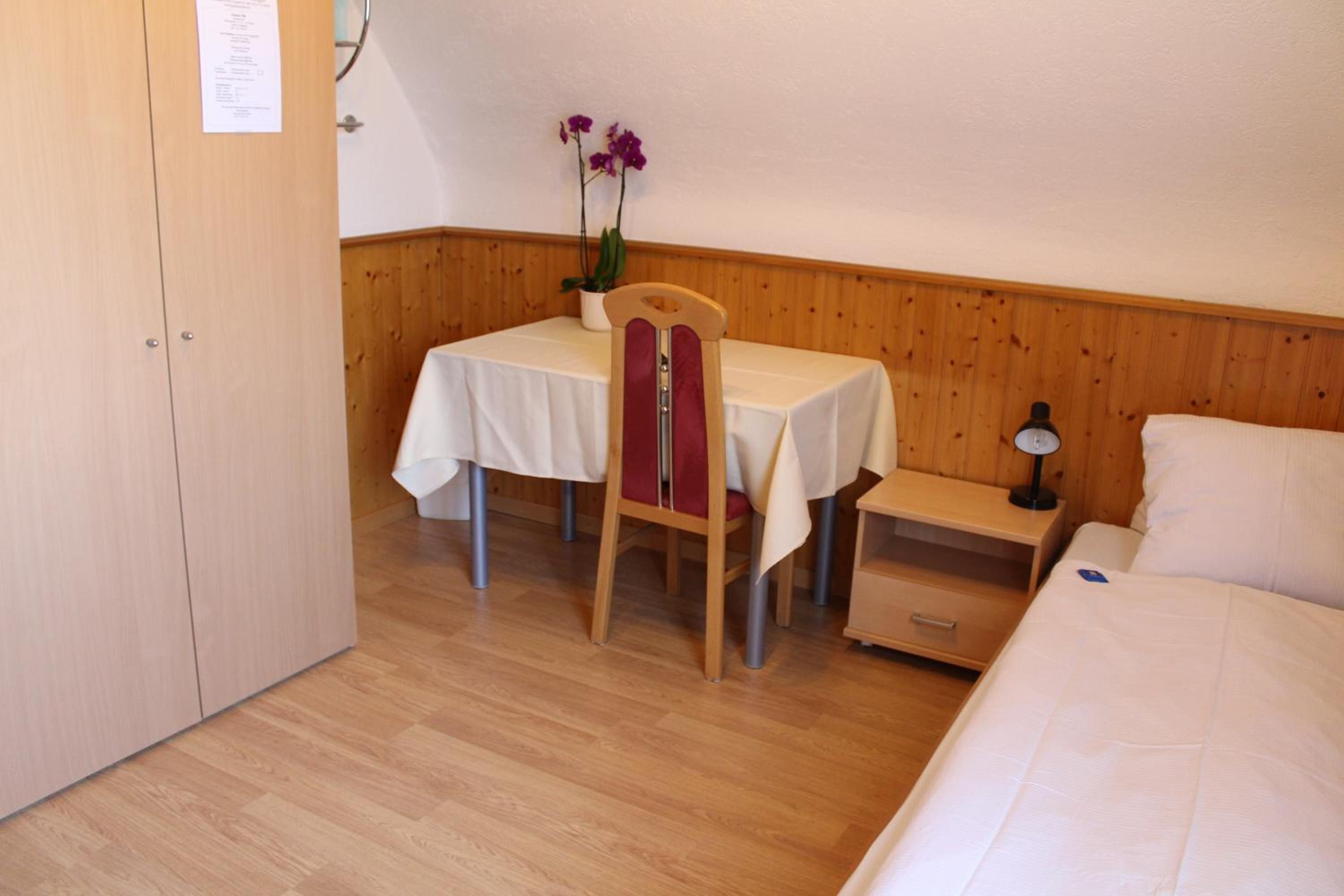 A room with a wooden floor, a bed, a table with a white tablecloth, a chair with a red cushion, a nightstand with a lamp, and a potted plant with purple flowers.