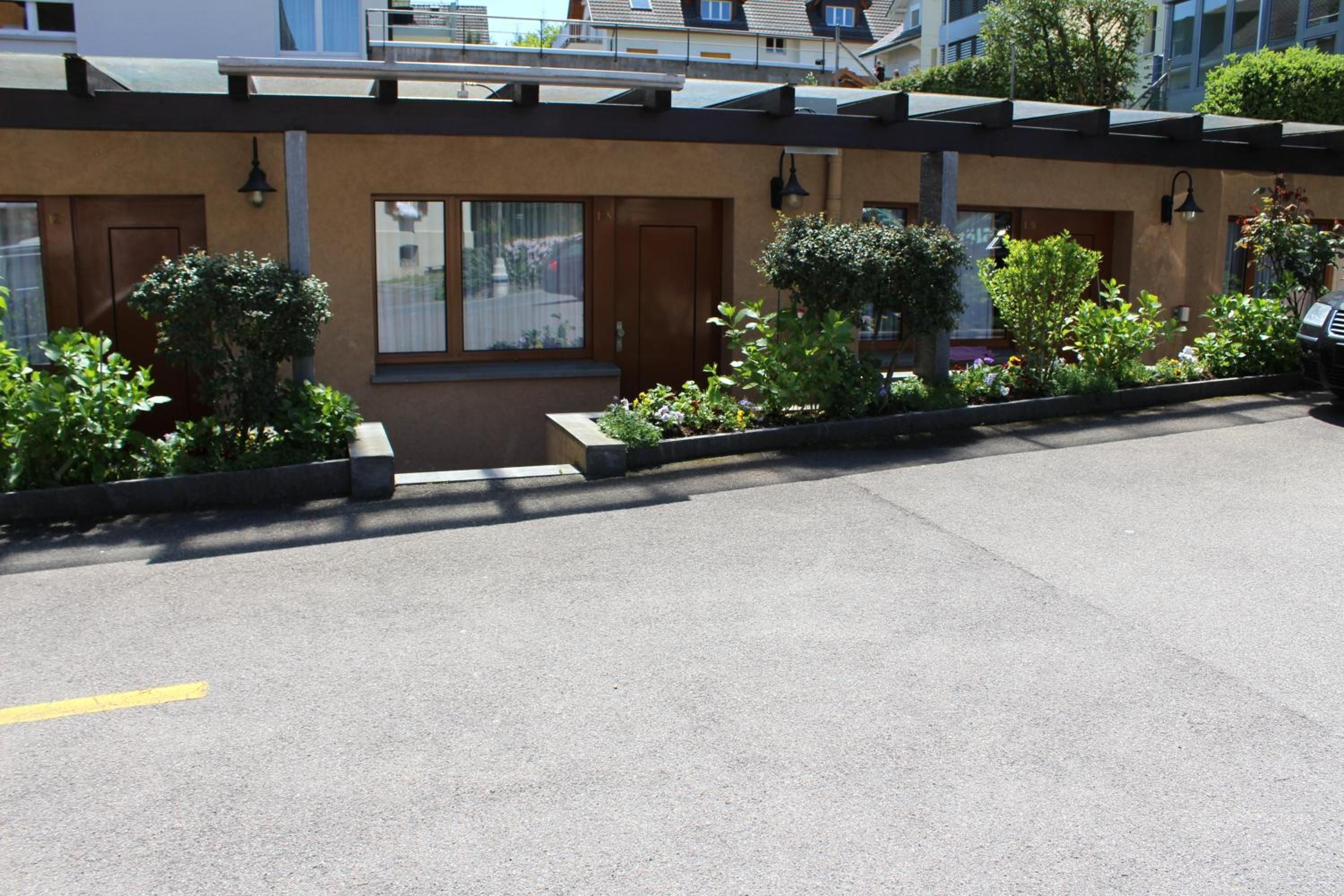 A parking area with a building entrance, a small garden with shrubs, and a clear sky.