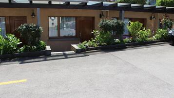 A parking area with a building entrance, a small garden with shrubs, and a clear sky.