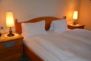 Cama doble con ropa blanca, dos lámparas de noche, un cabecero de madera y una mesita de noche con cajón.
