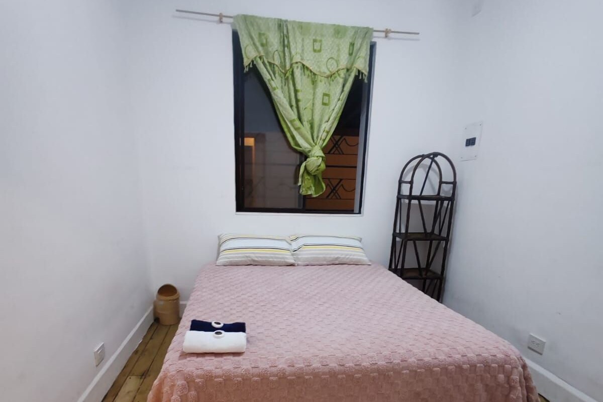Ein kleines Zimmer mit einem Einzelbett, Holzboden, einem grünen Vorhang und einem Metallregal.