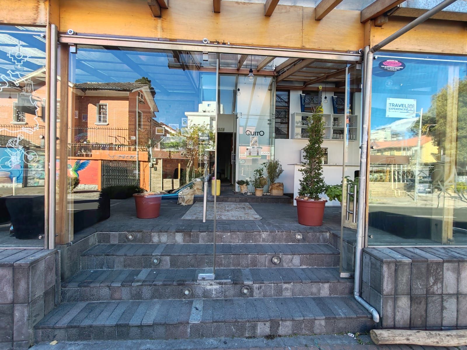 Una entrada de edificio con puerta de vidrio, escalones que la preceden y plantas en macetas a ambos lados.