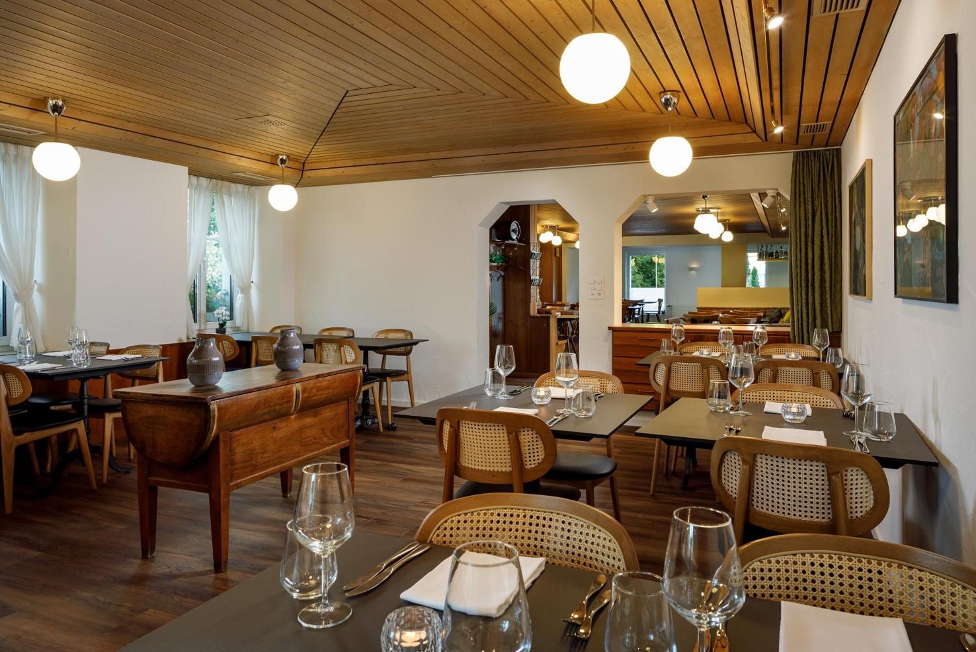 A restaurant with wooden furniture, round tables, and hanging pendant lights.