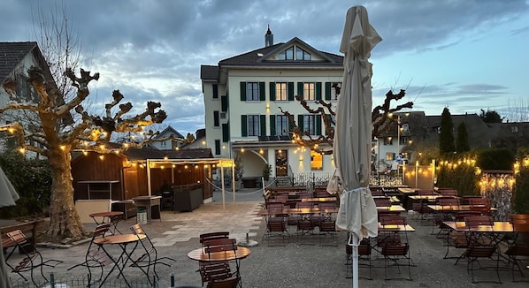 An outdoor seating area with tables and chairs, a building with green shutters, and string lights. - Hotel Lv Wen (Mönchaltorf)