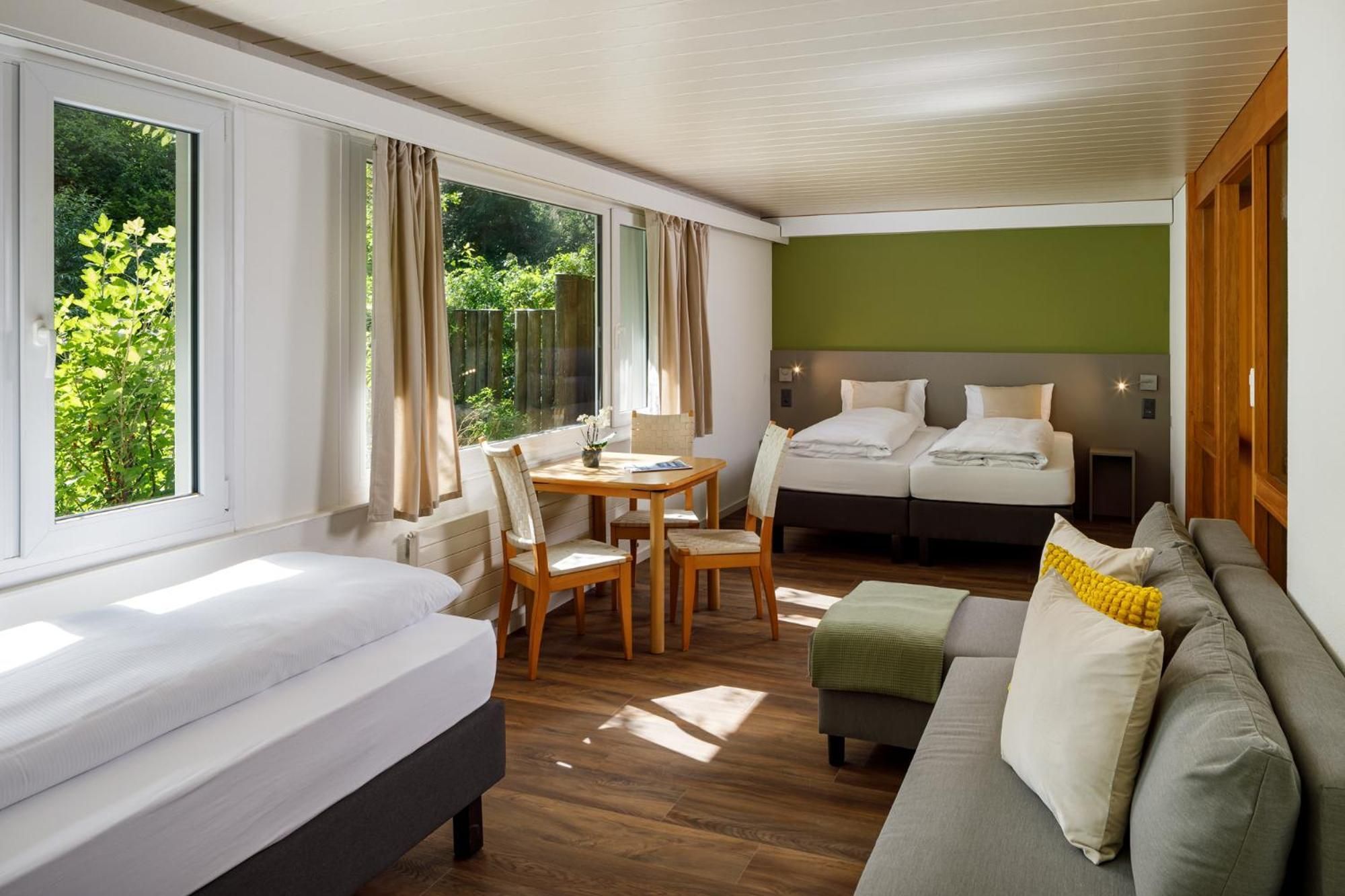 A hotel room with a bed, a sofa, a dining table with chairs, and a view of greenery through the windows.