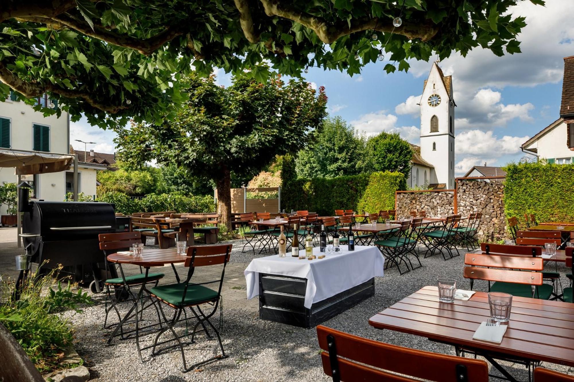 An outdoor dining area with tables and chairs, a barbecue grill, and a church in the background.