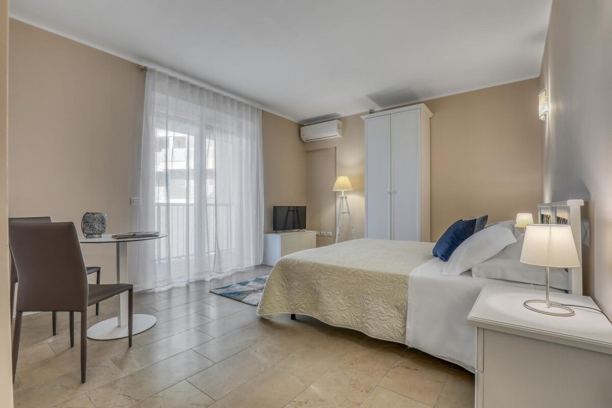 A hotel room with a bed, a desk, a chair, a TV, a lamp, and a window with curtains.