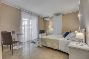 Habitación de hotel con cama, escritorio, silla, televisor, lámpara y ventana con cortinas.