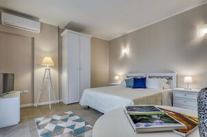 A hotel room with a bed, bedside lamps, a desk, a lamp, a magazine, and an air conditioning unit.