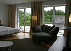 Une chambre moderne comprenant un lit, un canapé, un bureau et de grandes fenêtres donnant sur un paysage verdoyant.