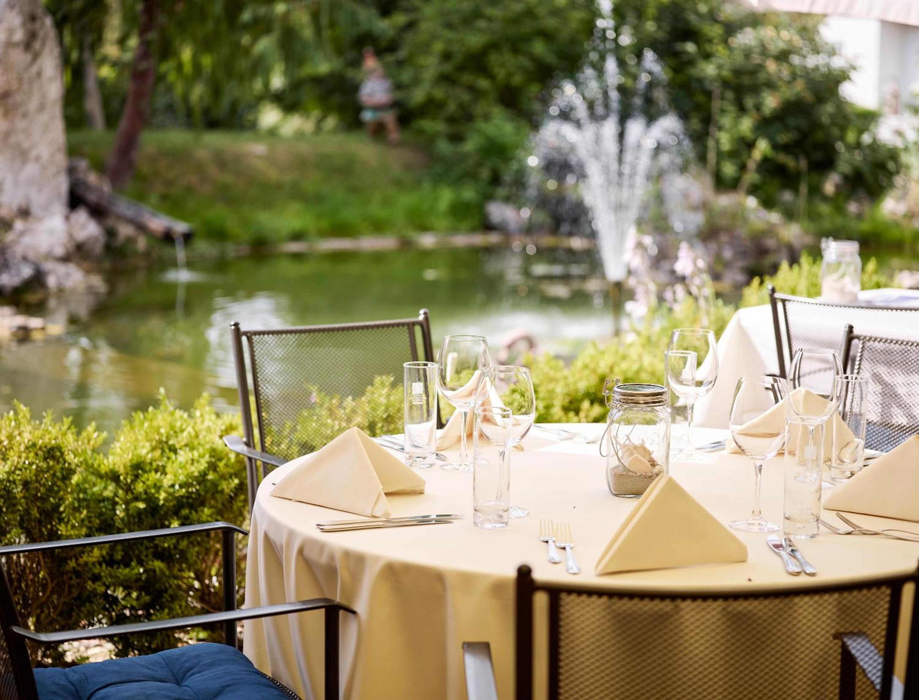 An outdoor dining area with round tables set for two, each with a white tablecloth, glassware, and cutlery.